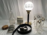 Photo showing bar table lamp with brass base and frosted white globe, crystal decanter, ceramic cognac dispenser, beer glasses, Levi's flask belt, and coaster or container lids