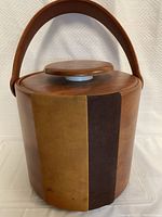 Front left angled view of the round ice bucket with brown and tan vertical suede or faux suede panels, showing the leather handle and lid knob.
