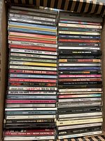 Photo shows a box filled with multiple music CDs in jewel cases stacked tightly. Visible CD titles include artists such as Beck, Big Shiny Tunes, and others.