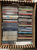 A cardboard box filled with multiple music CDs in jewel cases stacked horizontally and vertically showing spines and covers.