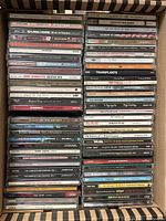 Photo showing a collection of approximately 50 music CDs stacked upright in a box, visible artist names include Sublime, U2, Bruce Springsteen, Talking Heads, Train, and others.