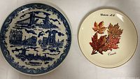 Blue and white decorative plate featuring Tower Bridge and other London landmarks next to cream decorative plate with autumn leaves and Canada text