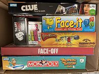 Top view of stacked board games showing Clue, Face-it, Villa Paletti, Face-Off, and Monopoly Tropical Tycoon boxes