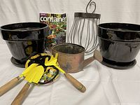 Full view of two black glazed terracotta pots, one with a chip on the bottom rim, metal watering can, gardening tool pouch with three yellow-painted metal tools with wooden handles, and Container Gardening book.
