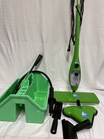 Photo showing green steam mop with triangular mop head and multiple black cleaning attachments laid out near a green plastic container for storage.