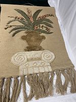 Full view of vintage jute wall hanging showing vase with flowers on a white pedestal and jute fiber fringe