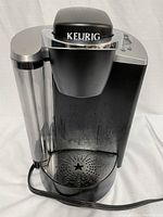 Front view of black and silver Keurig coffee maker with transparent water reservoir showing drip tray with star-shaped holes and Keurig logo on lid.