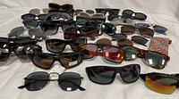 Overview photo showing over 20 pairs of sunglasses laid out on white surface, featuring multiple brands and styles.