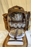Picnic backpack open showing secured utensils, 4 plastic glasses, 4 white plates, and plaid napkins.