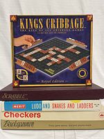Photo of boxed Kings Cribbage Royal Edition game on top of a stack of other boxed games: Scrabble, Merit Ludo and Snakes and Ladders, Checkers, and Backgammon