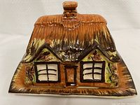 Front view of the ceramic cottage shaped covered butter dish, showing detailed painted roof, windows, and foliage with a chimney on top.