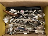 Top-down view of assorted vintage silverware in a box showing variety of spoons, knives, and forks with different handle designs