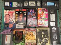 13 VHS tapes displayed flat, showing a mix of movie cover art and tape labels, including South Park, WWF sealed tape, Spaceballs, Life of Brian, and others.