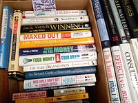 Photo showing a packed box with various business, investment, and success books arranged horizontally and vertically, titles visible including Winning by Jack Welch, Maxed Out, and others.