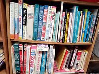 Photo showing two wooden bookcase shelves filled with more than 50 books about fitness, nutrition, yoga, meditation, tai chi, and diet, variety of colorful covers, some titles readable.