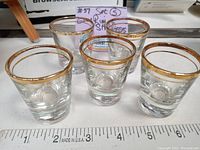 Five clear whiskey shot glasses arranged with a measurement ruler showing approximately 2.25 inches height. Each glass has a gold rim.