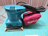 Side view of the blue Makita finishing sander with maroon dust bag attached, showing the power cord coiled on top.