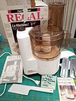 Full view of the Regal La Machine I food processor with translucent brown work bowl and feed chute pusher, original box behind, instruction manual, spatula and attachments laid out.