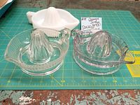 Top-down view of three vintage citrus juicers: two clear glass Sunkist juicers with handles and one white milk glass juicer in background.