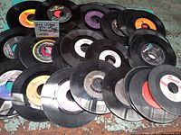 Photo showing a spread of many vintage 45 RPM records with various colored center labels, partially overlapping each other on a blue surface.