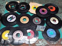 Photo showing 15 vintage 45 RPM vinyl records stacked and spread out, some in original paper sleeves with visible wear, displaying multiple label colors and record markings.