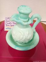 Front angle view of the aqua swirl milk glass pitcher/cruet with rose stopper sitting inside the matching bowl, showing embossed floral design and handle detail.