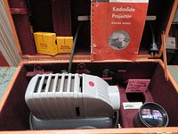 Photo of the slide projector in open pink leather-trimmed carrying case showing projector, accessories, instruction manual, bulb, power cord.