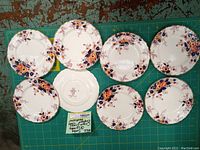 Eight 6-inch crown ducal plates with hand-colored floral patterns arranged on a green cutting mat, one plate flipped to show the maker's mark.