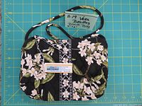 Full view of the vintage Vera Bradley handbag placed on a measuring grid, showing its size and black fabric with white floral prints and green leaves.