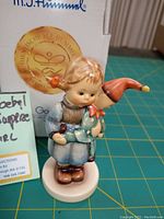 Front view of the M.J. Hummel porcelain figurine showing girl in blue dress holding elf doll with original box in background