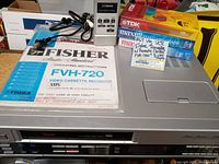 Fisher FVH-720 VHS Video Cassette Recorder with remote, manual, and tapes arranged on tabletop showing front tape-loading door, manual with blue and white cover, remote control with numeric keys, and 3 VHS tapes.