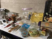 Wide view of lot with vintage tins, glass candy dish, Stanley sleeve bands, small wooden container, and other miscellaneous small collectibles spread on table.