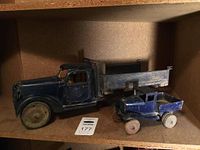 Two antique Buddy L toy trucks displayed side by side on a shelf, one larger black dump truck with a wooden bed and one smaller blue truck.