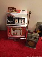 Both slot machine coin banks displayed side by side on a red surface.