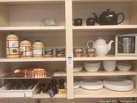 Wide shelf view showing dishes, cutlery tray, copper pans, canisters, mugs, and both black and white ceramic teapots