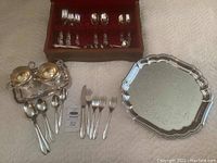 Full lot overview showing assorted silver plate flatware pieces, cream and sugar set, two engraved trays, and a box with extra flatware pieces.