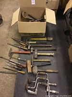 Photo capturing the array of vintage hand tools including hand drills, hammers, mallets, chisels, files, nail puller, and measuring rules spread on the ground and a cardboard box with extra tools.