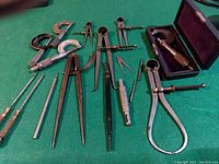 Collection of antique machinist precision measuring tools including micrometers, calipers, scriber, screwdrivers, cutter, punch, and drill bit starter laid out on green surface.