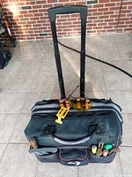 Front view of the Husky rolling tool bag with extendable handle pulled up, showing exterior pockets with visible hand tools such as chisels, tape measure, and safety glasses.