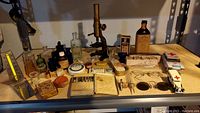 Overview photo showing the full collection of antique medical items including glass bottles, medicine tins, a brass microscope, boxes, vintage glasses and toy ambulances on a beige tabletop under shelving.