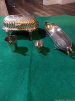 Photo showing silver plated butter dish with lid, footed dish with etched glass insert, and two sizes of silver plated jiggers on green cloth surface.