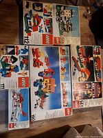 Photo showing instruction manuals for LEGO sets 112, 585, 111, 145, 190, and 770 arranged on a wooden table, partly overlapping, displaying images of built models and set numbers.