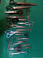Top view showing 20 assorted antique and vintage scissors laid flat on green cloth, varying sizes and handle styles from 4" to 14".