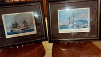 Two framed lithographs together on a wooden chair, showing maritime scenes with historic sailing ships and captions below each print.