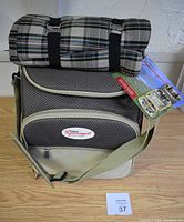 Photo of the closed cooler bag with the folded plaid picnic blanket secured on top with black straps, showing brand logo and carrying strap.