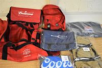 Overview of assorted bags showing red Canada 150 duffle bag, red Budweiser duffle and messenger bags, red and black Canada 150 backpack, blue Bud Light messenger bag, and gray Coors Light foldable bag.