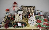 Full view of Christmas decorations including plush toys, wreaths, brass candle holders, ceramic candle holders, vintage light-up porcelain tree, wrapping paper, and Christmas cards arranged on surface.