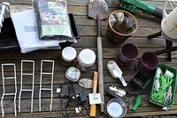 Photo showing a variety of garden tools, fertilizer containers, plastic pots, a hand trowel, pruning shears, ropes, and assorted small garden items arranged on a wooden surface.