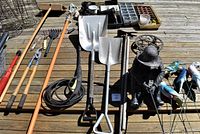 Photo showing garden hand tools, string trimmer, metal shovels, rake, black garden hose nozzle, seedling trays, decorative figurines including fisherman statue and colorful birds.