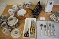 Overview of kitchen items on table including electric can opener, cutlery, plates, mugs, cutting boards, and utensil holder.